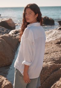 Camicia bianca leggera con motivo strutturato, maniche arrotolate e vestibilità comoda, abbinata a un denim azzurro. Oceano e rocce sullo sfondo.