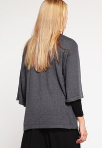 Woman with long blonde hair wearing a loose dark gray short-sleeve top over a black long-sleeve shirt, facing away against a white background.