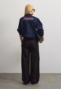 Donna bionda con giacca di jeans scura con logo "pegador" sulla schiena, pantaloni neri a gamba larga, occhiali da sole, che tiene una pochette nera, di spalle.