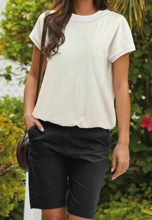 Kvinde iført en hvid kortærmet bluse med blondekant og sorte shorts, stående udendørs med grønne planter og blomster i baggrunden.