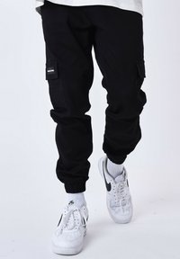 Pantaloni cargo neri con polsini elasticizzati, dotati di due tasche laterali e una piccola patch. Abbinati a scarpe da ginnastica bianche e calzini bianchi.