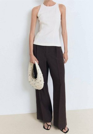 Vrouw draagt mouwloos wit topje en wijde donkere broek, houdt een wit halvemaanvormige handtas met studs vast, staat op blote voeten.