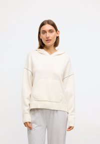 Cream hooded sweatshirt with a front pocket, soft texture, ribbed hem, and sleeves. Styled with gray sweatpants.