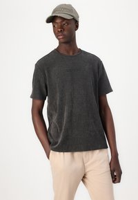 T-shirt à manches courtes gris foncé avec le logo "GUESS" brodé. Porté avec un pantalon beige et une casquette gris clair. Texture lisse, coupe décontractée.