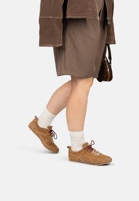 Sneaker in suede marrone con lacci bordeaux, dettagli testurizzati e design a profilo basso, abbinati a calzini bianchi alla caviglia e un outfit marrone.