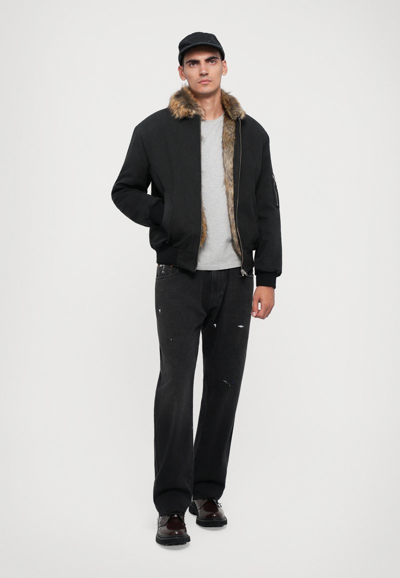 Black bomber jacket with fur collar, gray t-shirt, black distressed jeans, and dark shoes; includes zipper and side pocket detail.