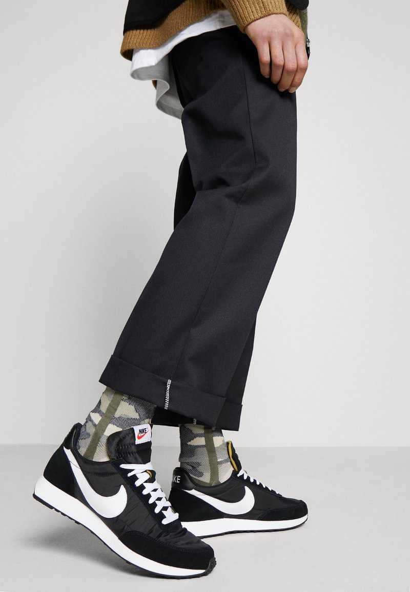 Personne portant des baskets Nike noires et blanches avec des chaussettes camouflage et un pantalon noir court debout sur un sol gris.