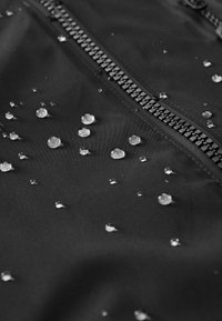 Tissu imperméable noir avec une fermeture éclair, présentant des gouttelettes d'eau éparses reflétant la lumière. Texture lisse avec une finition mate.