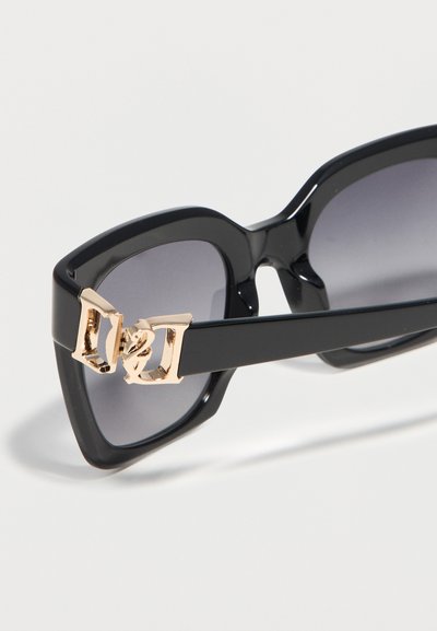 Μαύρα γυαλιά ηλίου σε σχήμα γάτας με σκούρους φακούς και χρυσή διακοσμητική άρθρωση στο μπράτσο του σκελετού, πάνω σε λευκό φόντο.