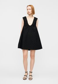 Femme portant une robe noire sans manches avec un col en V profond et un tissu ajouré, associée à des sandales noires à bride autour de la cheville, debout devant un fond blanc.