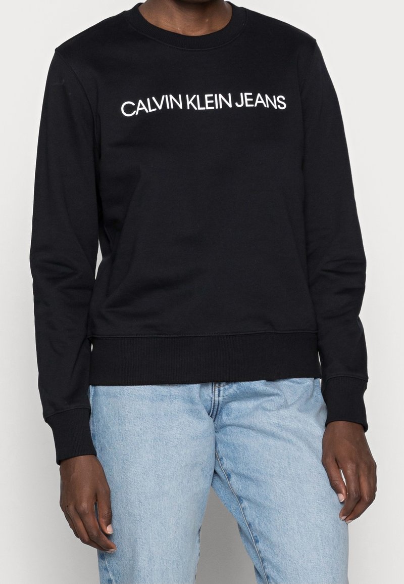 Černá mikina s bílým logem "CALVIN KLEIN JEANS", kulatý výstřih, ribbenková manžeta a lem, vyrobená z měkké tkaniny. Nošena s světlými modrými džíny.