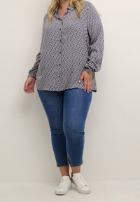 Personne aux courbes portant une blouse boutonnée à motifs bleus, un jean skinny bleu cropped avec fente à l'avant et des baskets blanches, se tenant contre un fond uni.