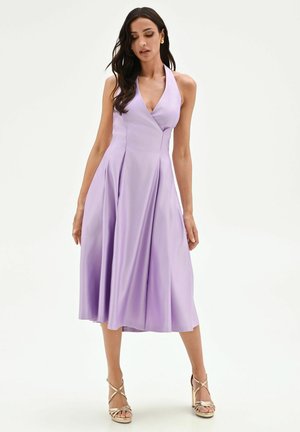 Femme portant une robe midi lavande sans manches à col halter en V avec une jupe plissée, associée à des sandales à talons hauts dorées et à brides.