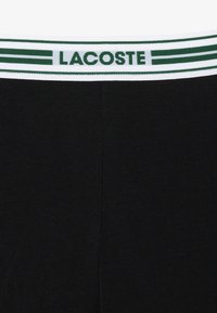 Tessuto a costine nero con un waistband a righe bianche e verdi che presenta "LACOSTE" in testo verde. Design semplice e funzionale.