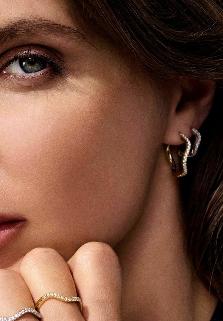Guld- och silverörhängen med en vågig design, prydda med klara ädelstenar. Ringarna på fingrarna är böjda med accentstenar, vilket visar eleganta konturer.