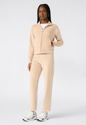Felpa beige con zip e strisce bianche di accento, abbinata a pantaloni affusolati coordinati. Indossata con sneakers bianche, dotate di accenti neri.