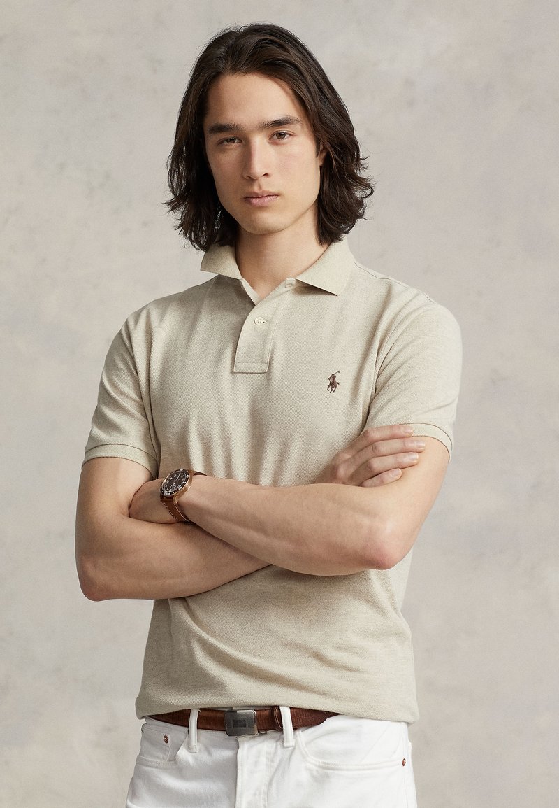 Béžové polo tričko s krátkym rukávom a golierom, s malým logom. Model má dlhé vlasy a nosí hnedé hodinky s koženým remienkom.