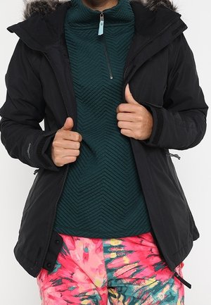 Person iført en sort vinterjakke med pelskrave, mørkegrøn tekstureret pullover og farverige bukser med mønstre i pink, grøn og gul.