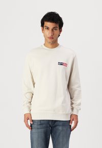Kremfarget bomullssweatshirt med lange ermer, ribbede mansjetter og kant. Har et farget logo-patch på venstre bryst. Bæreres med blå jeans.