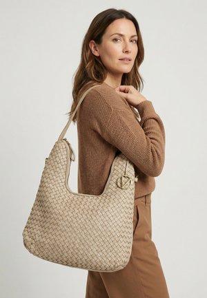 Kvinde iført en brun sweater og bukser holder en stor beige flet skuldertaske med en gylden rund spænde.