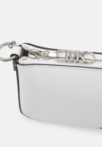Bijela kožna torbica s srebrnim zatvaračem, s privjeskom sa logotipom "MK". Rubovi su obrubljeni smeđom bojom, stvarajući kontrast duž šavova.