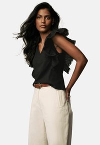 Top a spalline nere con volant e scollatura a V, realizzato in tessuto testurizzato e rifinito con dettagli decorativi, abbinato a pantaloni beige a gamba larga e una cintura marrone.