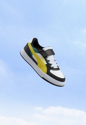 Baskets Puma noires, blanches et jaunes avec lacets et bande Velcro flottant sur un fond de ciel bleu clair.