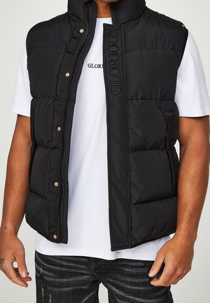 Mann trägt schwarze gesteppte Weste mit der Aufschrift "GLORIOUS" auf der Vorderseite über weißem T-Shirt und schwarzer Jeans.