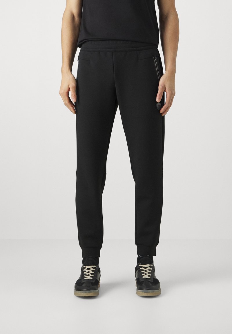 Svarta tapered sweatpants med en mjuk textur, elastisk midja, sidofickor med dragkedja och ribbad mudd, bärs med svarta sneakers.