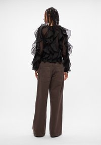 Blusa nera trasparente con strati a volant; abbinata a pantaloni larghi marroni. La blusa ha maniche lunghe e un collo alto.