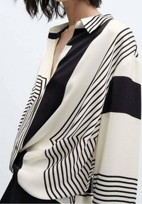 Chemise à rayures noires et blanches avec motifs géométriques, col et coupe décontractée. Le tissu semble lisse avec un léger éclat.