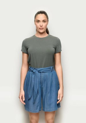 Frau mit dunklen Haaren im Pferdeschwanz, die ein grünes kurzärmliges Shirt und hoch taillierte blaue Jeans-Shorts mit gebundenem Gürtel trägt, vor schlichtem Hintergrund stehend.