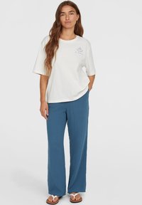 Weißes Baumwoll-T-Shirt mit kurzen Ärmeln und rundem Ausschnitt, kombiniert mit blauen strukturierten, weit geschnittenen Hosen und Flip-Flops.