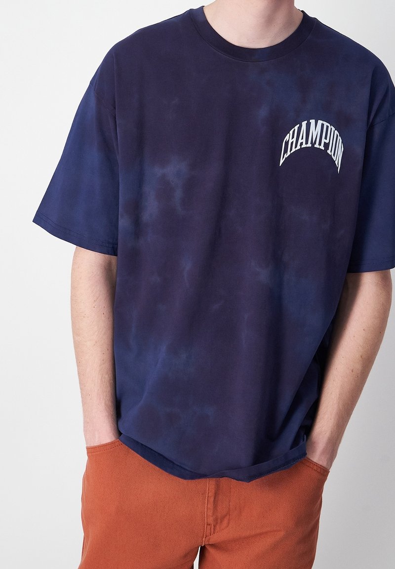 Camiseta de algodón de tie-dye en color azul marino de manga corta, con un logo blanco de "CHAMPION" impreso en la parte superior del lado izquierdo del pecho. Textura suave.