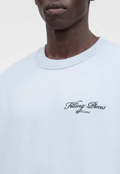 Gros plan d'une personne portant un sweat-shirt bleu clair avec "Filling Pieces" et un petit texte brodé sur la poitrine.