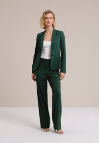 Žena sa kratkom plavom kosom nosi tamnozeleni sako i široke hlače u istoj boji, bijelu majicu i sandale na petu.