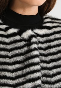 Garment in pelliccia artificiale a strisce bianche e nere con una texture morbida, caratterizzato da un collo alto e strisce verticali prominenti che alternano spessore.
