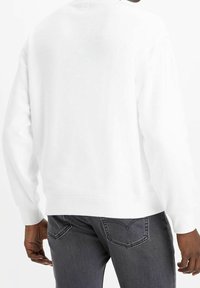 Felpa bianca in cotone con collo rotondo, maniche lunghe, polsini e orlo a coste. Indossata sopra jeans grigio scuro con tasche posteriori.