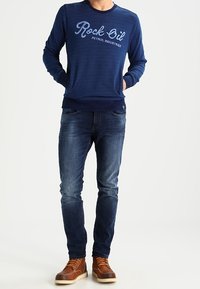 Námořnický svetr s kulatým výstřihem a bílým logem "Rock Oil", spojený s modrými džíny a hnědými šněrovacími botami. Pohodlný, přiléhavý design.