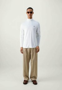 T-shirt branca de manga longa com gola alta, combinada com calças bege claras e sapatos castanhos. Acompanhada de óculos de sol escuros.