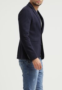 Navy blazer with a tailored fit, single button closure, notch lapels, and three-button cuffs, paired with light blue distressed jeans.