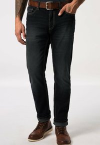 Jeans in denim blu scuro con cuciture gialle, slim fit, design a cinque tasche, indossati con una cintura in pelle marrone e stivaletti marroni con lacci.