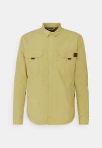 Camisa chaqueta de manga larga en color caqui claro con botón en la parte delantera, dos bolsillos de pecho con solapa y detalles en negro en las solapas de los bolsillos.