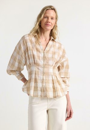 Kvinde med blondt hår iført beige og hvid ternet skjorte med knapper og offwhite bukser, stående mod en ensfarvet lys baggrund.