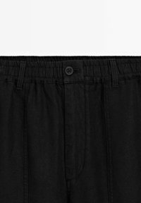 Pantalons noirs en tissu texturé, avec une taille élastique et un bouton, et des plis plats à l'avant. Aucune poche n'est visible sur l'image.