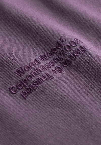 Lilla stoff med myk tekstur som har brodert tekst: "Wood Wood Copenhag 2002" og en falmet slagord, plassert fremtredende.
