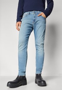 Jeans de denim azul claro con un ajuste slim, cinco bolsillos y un ligero desgastado. Llevados con botas negras hasta el tobillo con una suela gruesa.