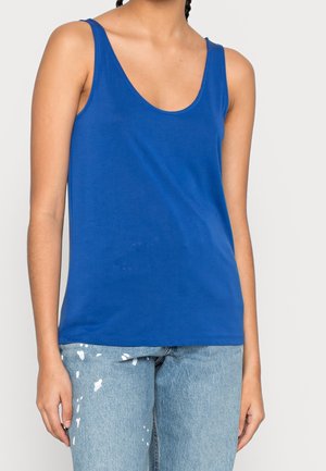 Débardeur bleu roi sans manches à encolure dégagée porté avec un jean en denim bleu clair éclaboussé de peinture, sur une personne debout.