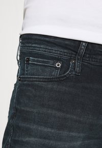 Os jeans de ganga escura apresentam um design ajustado com uma textura suave, costuras em contraste e rebites metálicos nos bolsos para um detalhe adicional.