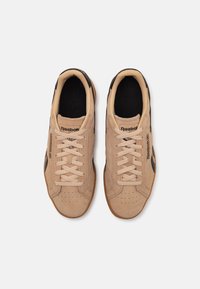 Baskets en daim beige avec des accents noirs, présentant des bouts ronds, des lacets plats, des perforations et une semelle en caoutchouc. Logo sur le côté.